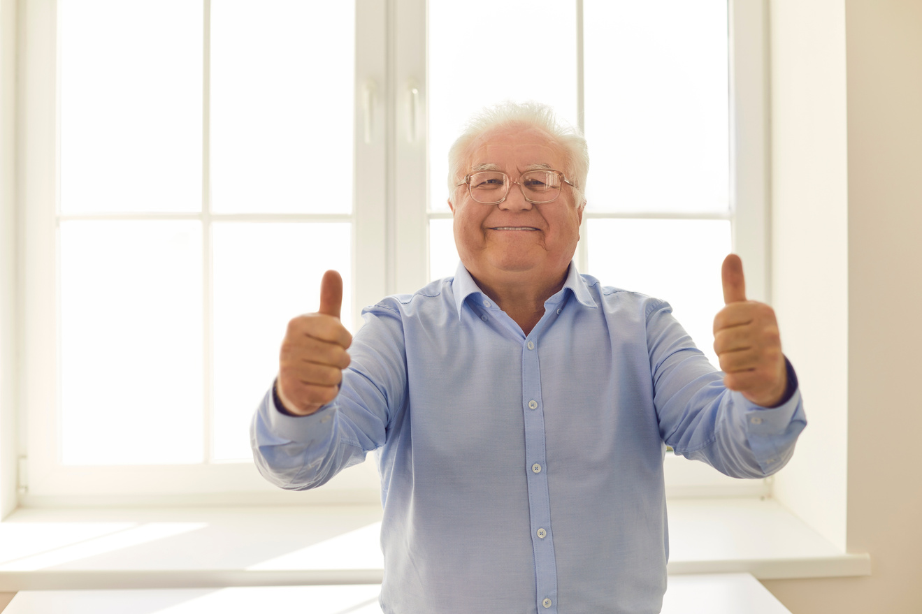 Happy Senior Man Giving Thumbs-up and Looking at Camera Standing near the Window